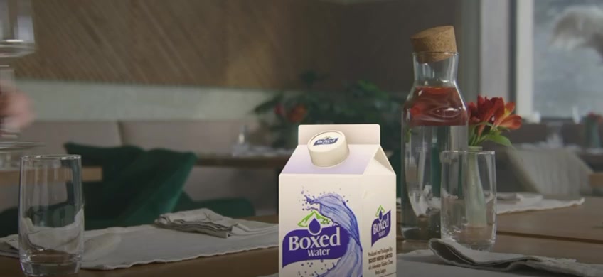 Boxed Water carton on a dining table beside drinking glasses and a water carafe.